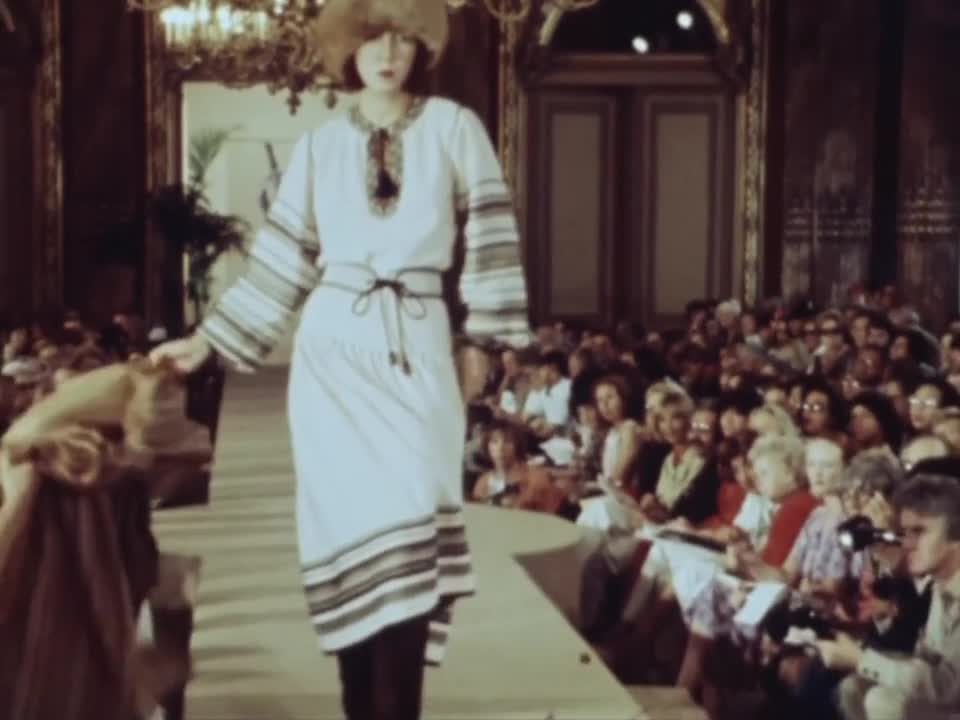 Film of the Yves Saint Laurent Autumn-Winter 1976 show at the Hôtel Inter-Continental: a model in a white peasant dress with striped bands walks down the runway