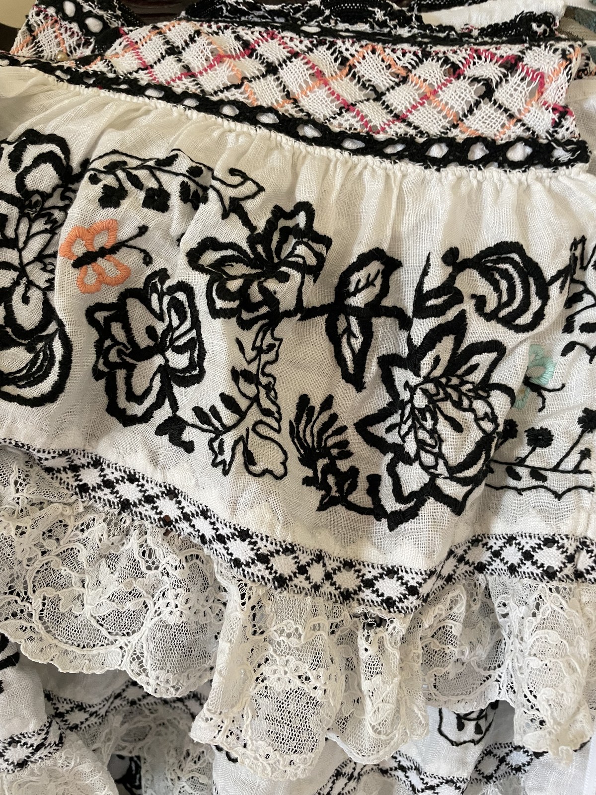 Close-up of a pollera: black embroidery on white fabric, a band of bobbin-lace braid over the seam between the tiers and lace below