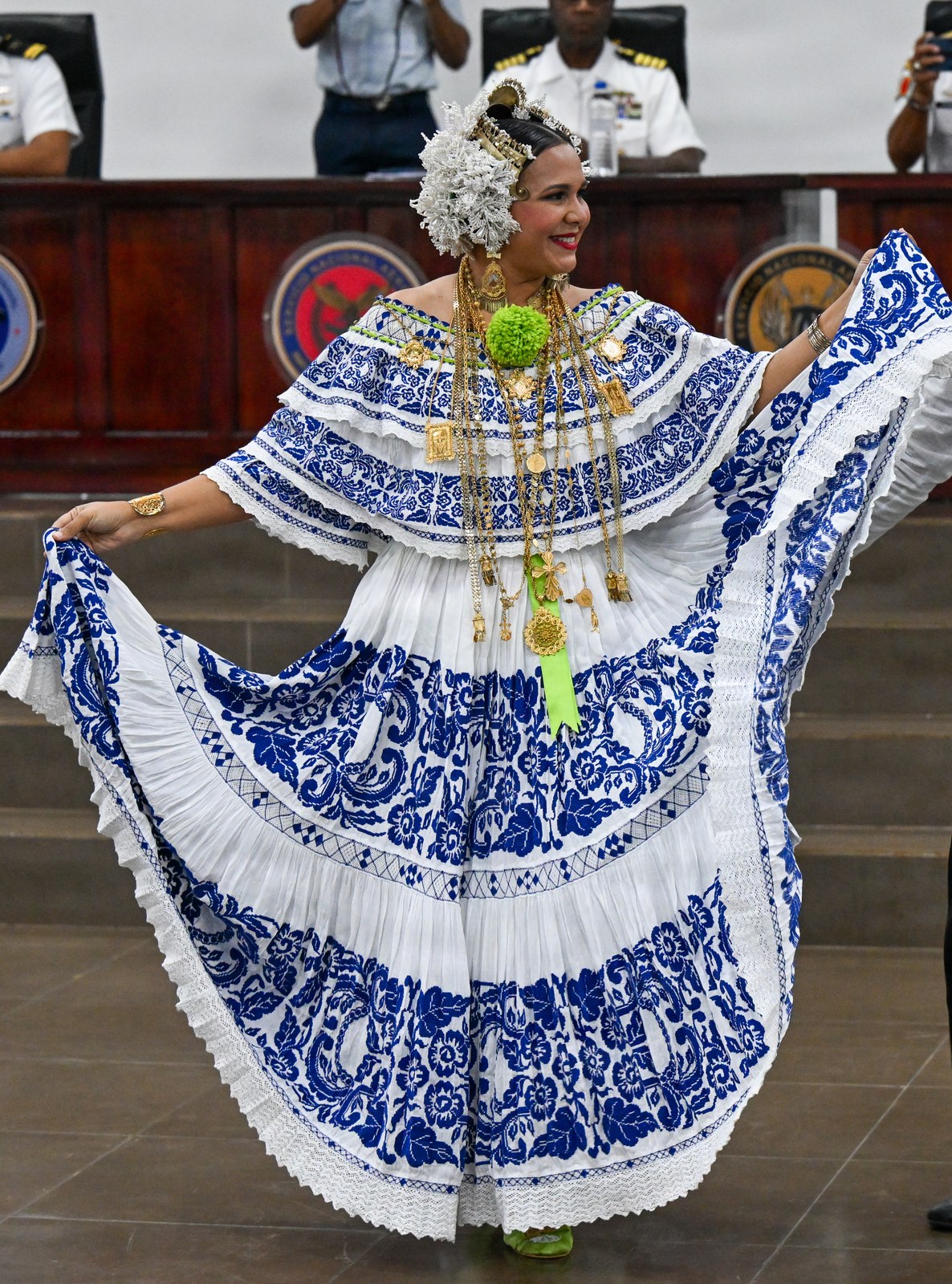 A dancer in a white pollera de gala embroidered in blue: a wide skirt of two gathered tiers with lace at the hem, gold chains and a headdress of tembleques