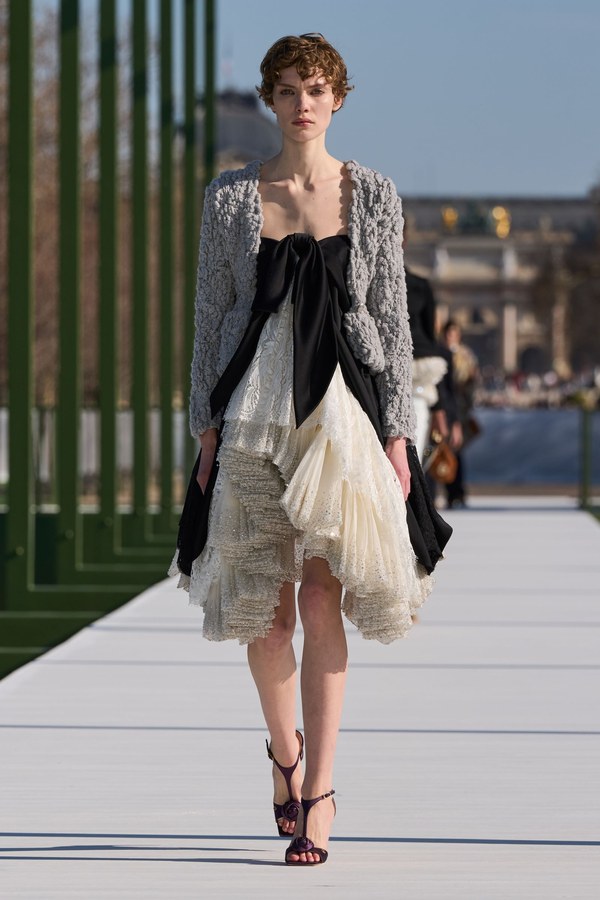 Dior Fall 2026, look 59: black overskirt open at the front over rows of ivory lace ruffles